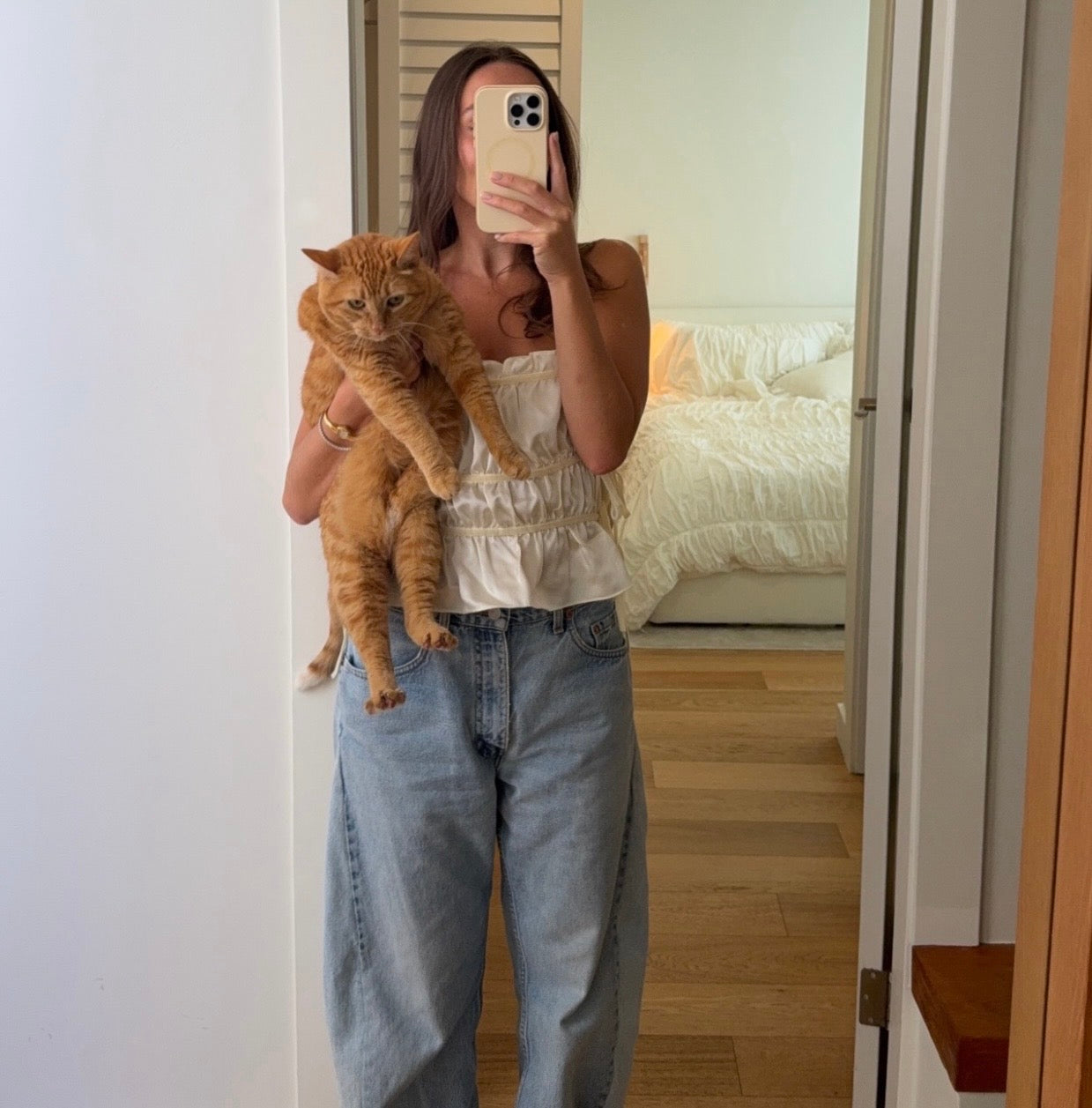 Person holding an orange cat in a room with a bed and wooden floor.